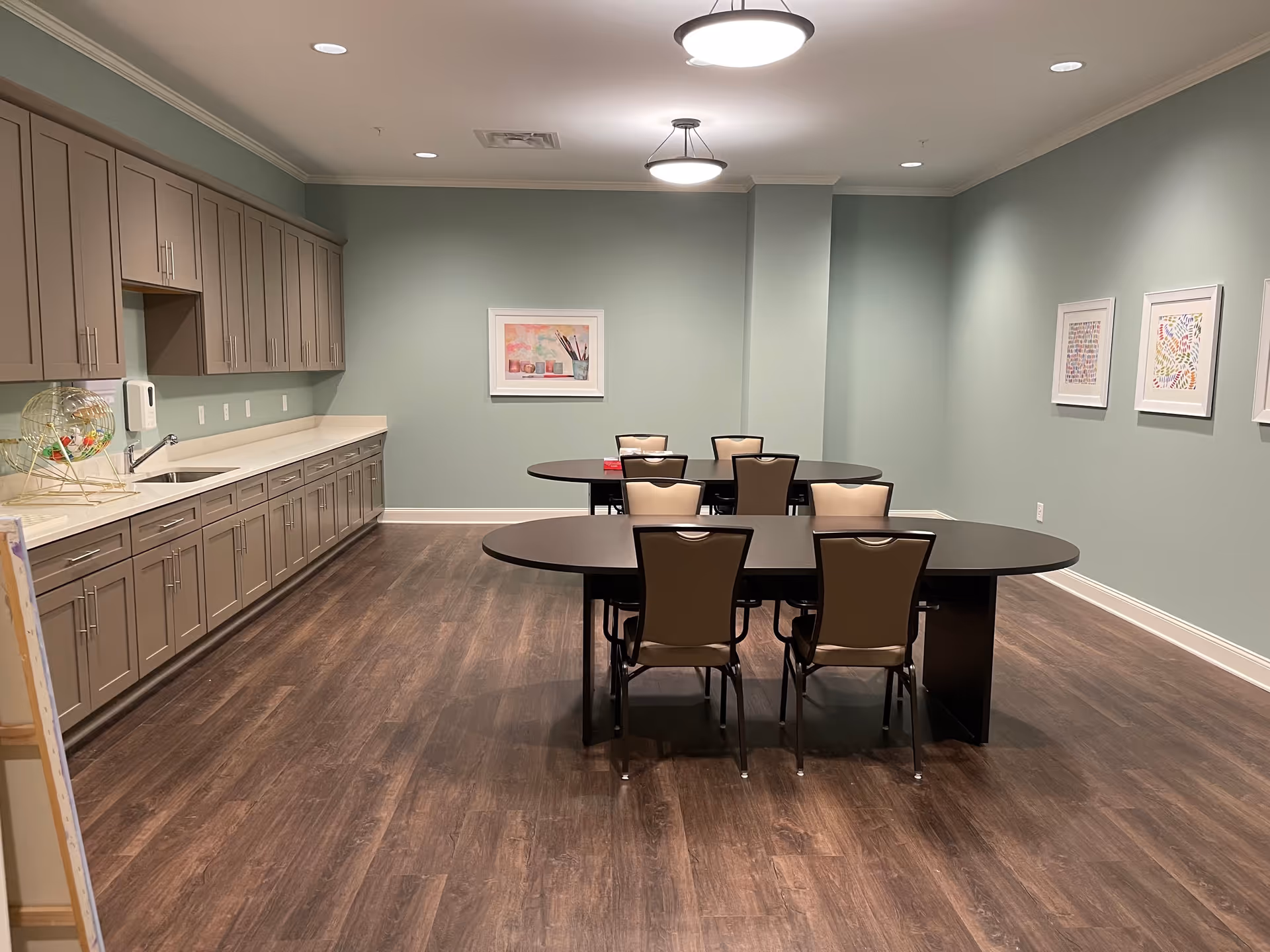 A spacious room with two oval tables and six chairs arranged around them. The room has light green walls, wooden flooring, and overhead circular light fixtures. On the left side, there is a long countertop with a sink and multiple cabinets above and below. The walls are decorated with framed artwork.