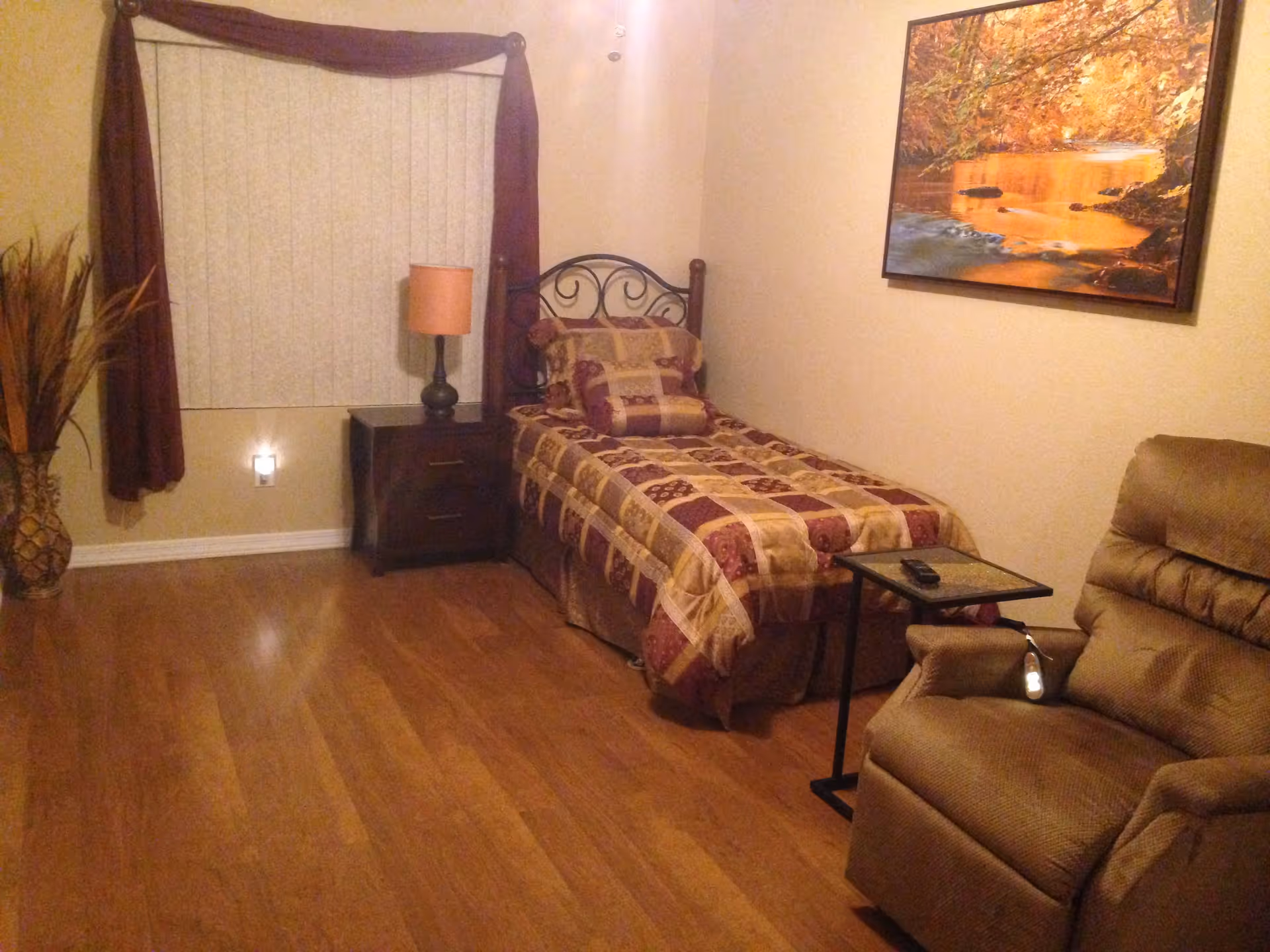A cozy bedroom with a single bed covered in a patterned quilt, a nightstand with a lamp, a reclining chair with a small side table holding a remote control, a window with vertical blinds and dark curtains, a decorative plant in a vase, and a framed picture of a serene autumn river scene on the wall.