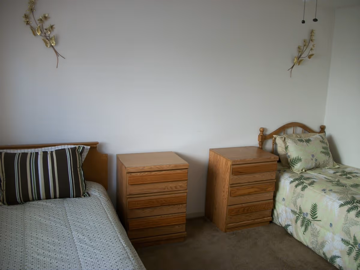 A simple bedroom with two single beds separated by two wooden nightstands. The bed on the left has a striped pillow and a light-colored bedspread, while the bed on the right has a pillow and bedspread with a green leaf pattern. The walls are plain white with two decorative wall hangings featuring gold butterflies and flowers.