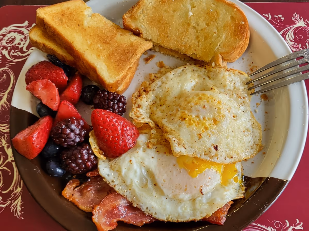 A plate of breakfast food including two fried eggs, two slices of toasted bread, several pieces of bacon, and a mix of fresh berries such as strawberries, blackberries, and blueberries on a decorative plate with a red and white patterned placemat underneath.