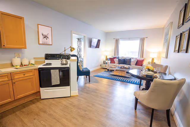 A bright and cozy living space featuring a kitchen area with wooden cabinets and a white stove on the left, a small dining table with two upholstered chairs on the right, and a living room area with a sofa, colorful cushions, a coffee table, and a wall-mounted TV. The room has light-colored walls, wood flooring, and a large window with curtains letting in natural light.