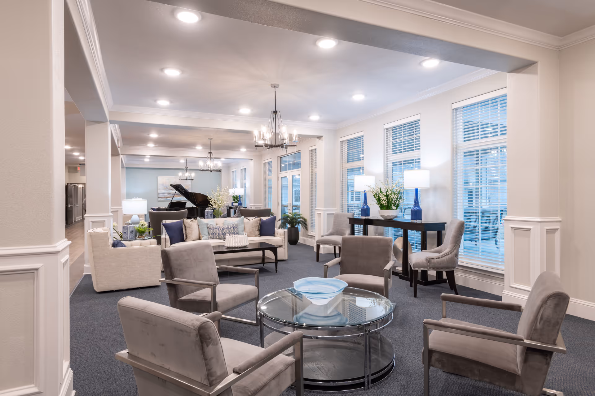 A spacious and well-lit living room area in Wesley House featuring multiple seating arrangements including four gray armchairs around a glass coffee table with a decorative bowl, a beige sofa with blue and patterned cushions, and additional chairs near windows with white blinds. The room has recessed ceiling lights, chandeliers, and table lamps on a console table with blue vases and floral arrangements. A grand piano is visible in the background.