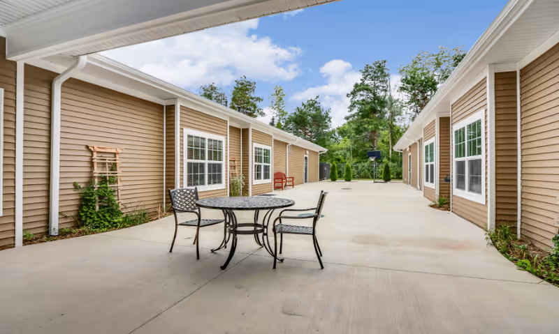 Outdoor courtyard area between two single-story buildings with beige siding and white trim. The courtyard has a concrete floor with a round metal table and three chairs in the center. There are some plants along the building walls and trees in the background under a partly cloudy sky.