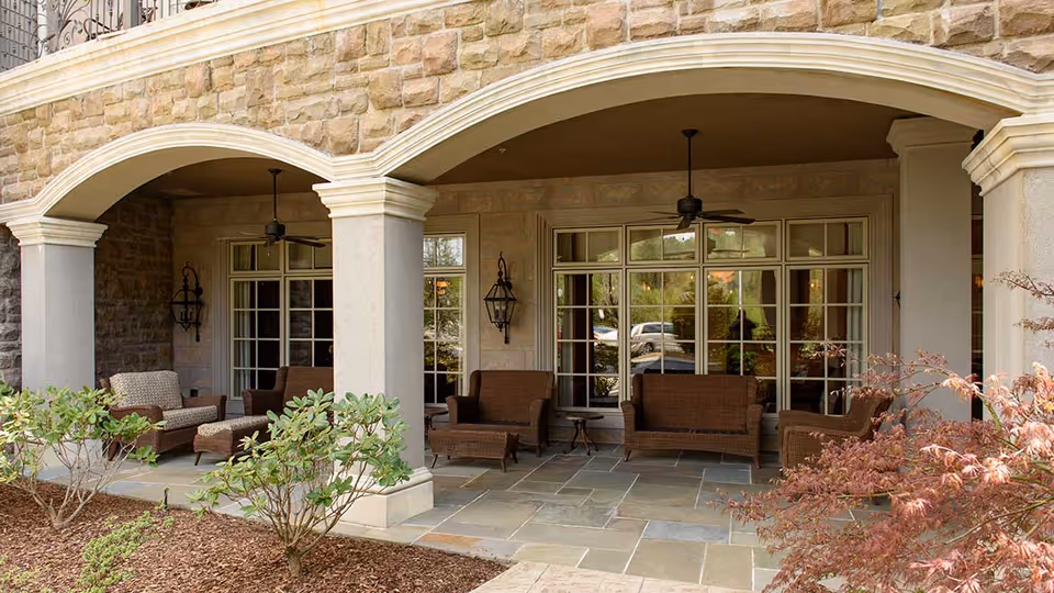 Covered outdoor patio area with stone flooring and stone pillars supporting arches. The patio is furnished with wicker seating including chairs and sofas with cushions, small tables, ceiling fans, and wall-mounted lantern-style lights. There are shrubs and plants in the foreground.