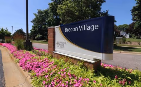 Blue Brecon Village sign with pink flowers along sidewalk by the road.