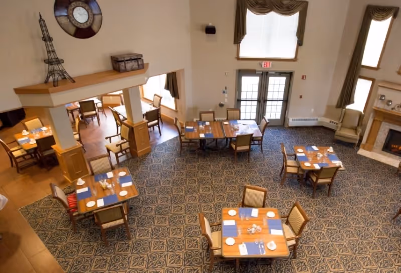 A dining room in a senior living facility with several wooden tables set with plates, napkins, and utensils. The room has patterned carpet flooring, large windows with drapes, a fireplace, and a clock on the wall. Chairs are arranged around the tables, and the space is well-lit with natural light.