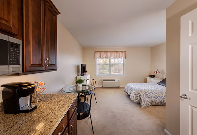 A senior living studio apartment featuring a kitchenette with granite countertops, a coffee maker, and a microwave. There is a small round glass table with two chairs near the window. The room has a bed with patterned bedding, a nightstand with a lamp, and a wall-mounted TV. The window has a valance and an air conditioning unit below it.