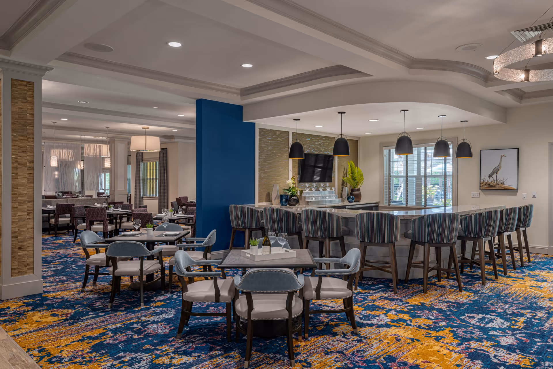 A spacious dining area in a senior living facility featuring multiple tables with chairs, a bar counter with high stools, pendant lights hanging from the ceiling, decorative plants, a wall-mounted TV, and large windows allowing natural light. The room has a colorful patterned carpet and a modern, welcoming design.