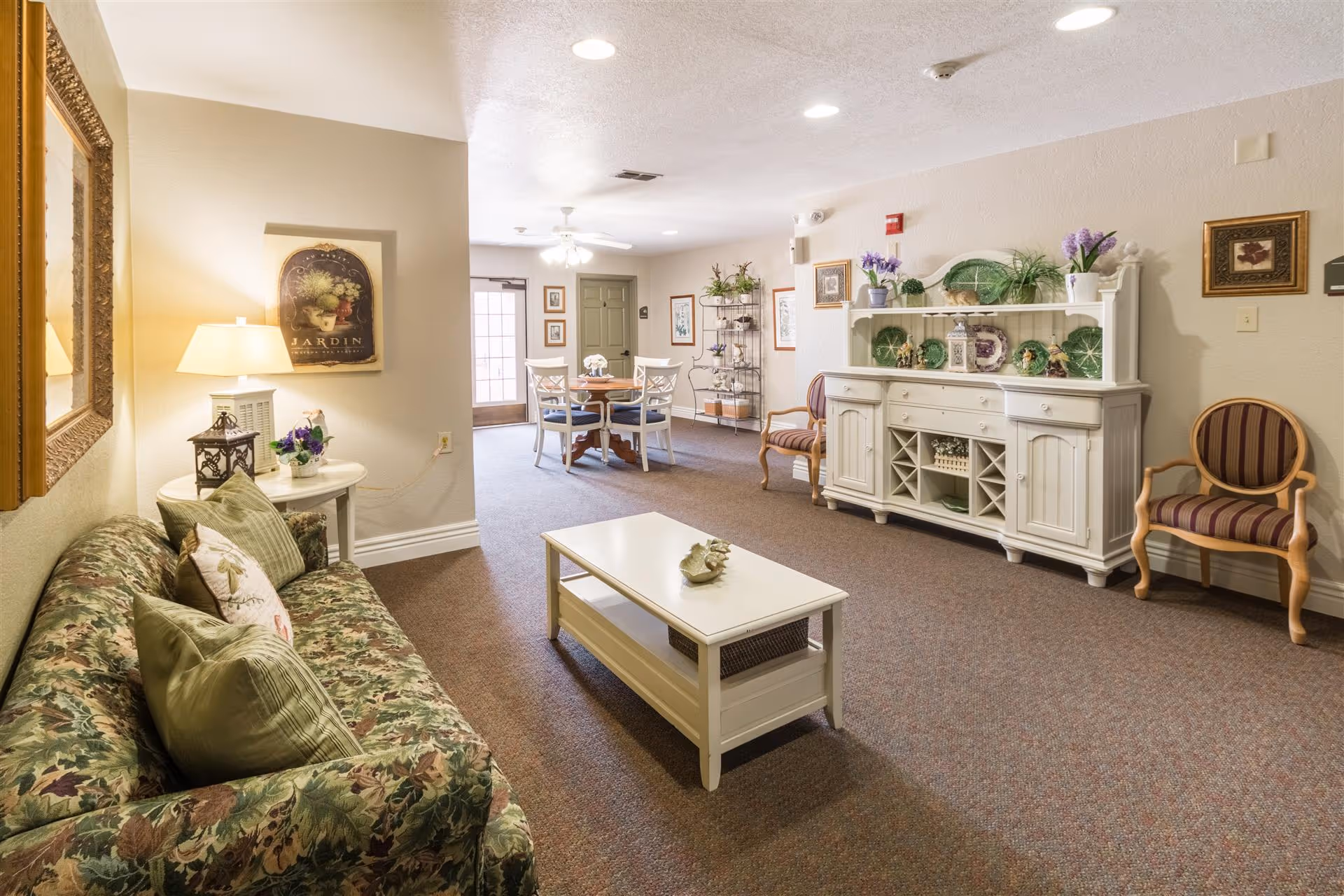 A bright assisted-living common room with a floral sofa, coffee table, white sideboard, and a dining table in the background.