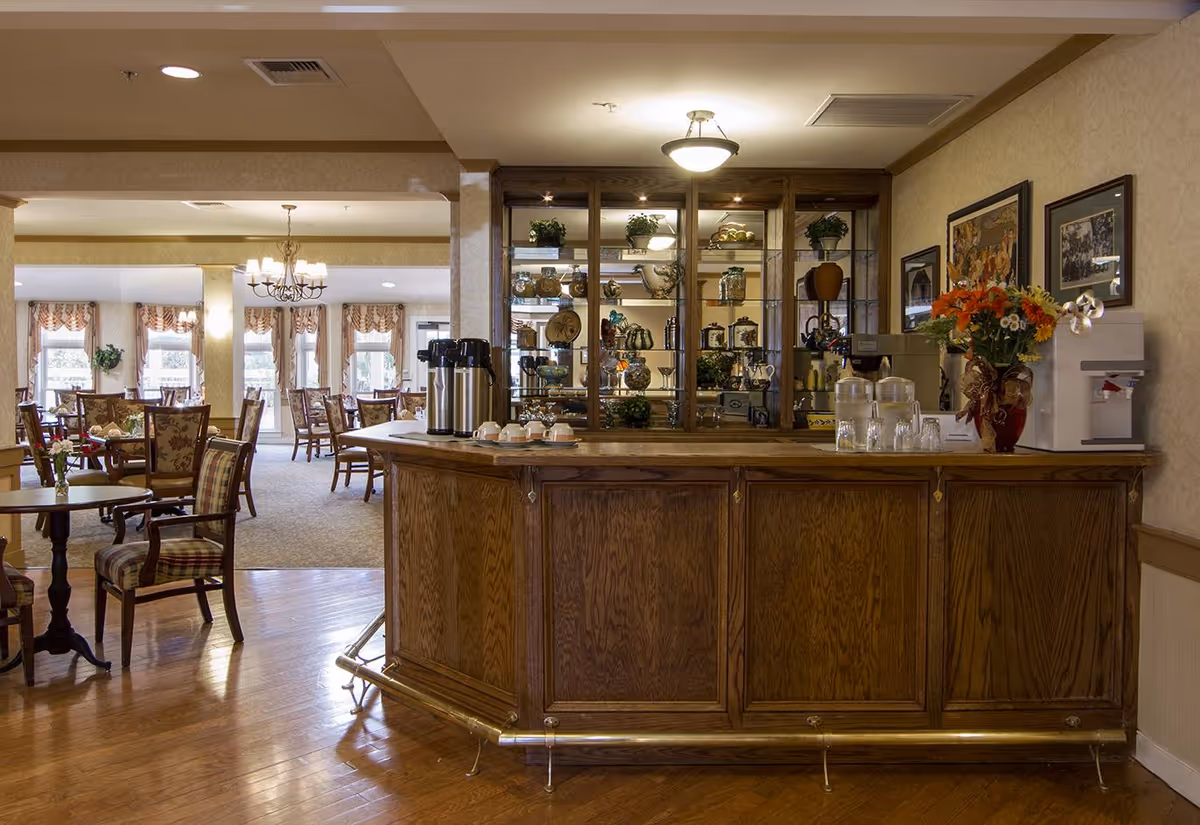 Wood-paneled service counter and coffee station in a senior living dining area with tables, chairs, and decorative shelving in the background.