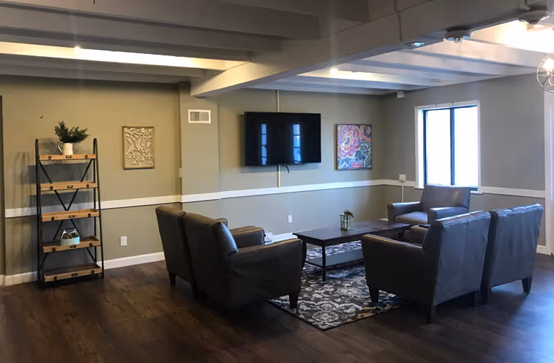 A cozy living room area with four dark leather armchairs arranged around a rectangular coffee table on a patterned rug. A flat-screen TV is mounted on the wall above the table. There is a window letting in natural light, a decorative shelf with plants and ornaments, and two pieces of wall art. The room has wooden flooring and neutral-colored walls with white trim.