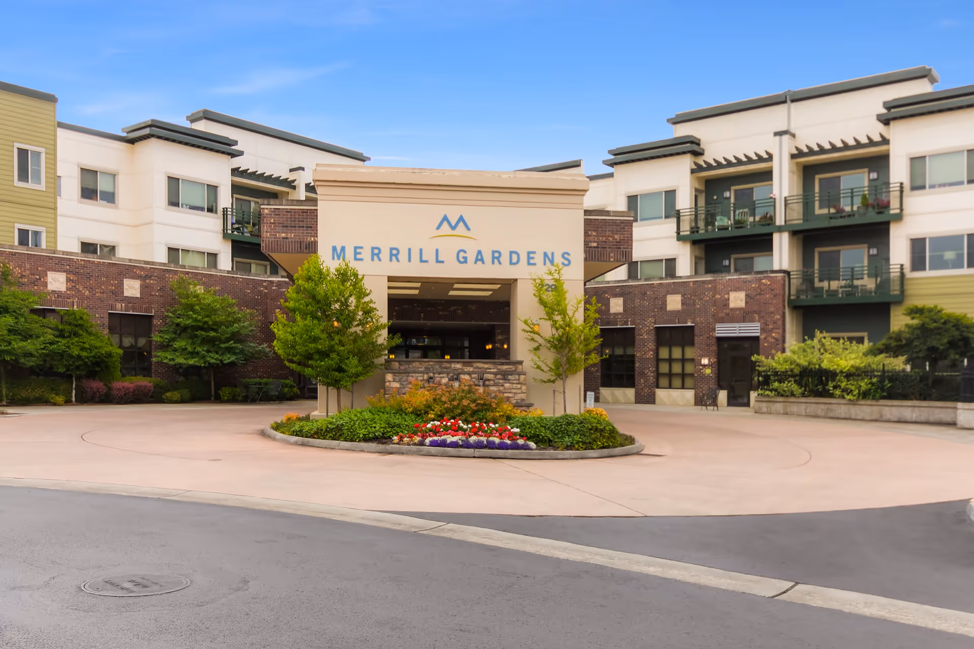Front exterior view of Merrill Gardens senior living facility showing a circular driveway with landscaped greenery and flowers in the center, surrounded by a multi-story building with balconies and large windows under a clear blue sky.