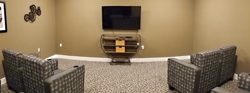 Small TV lounge with a wall-mounted flat-screen, two upholstered chairs, patterned carpet and neutral-colored walls.