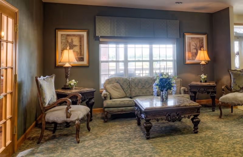 A cozy living room with classic wooden furniture including a cushioned sofa, two armchairs, and a carved wooden coffee table with a floral arrangement. Two side tables with lamps and framed botanical artwork are on either side of a large window with a valance, letting in natural light.