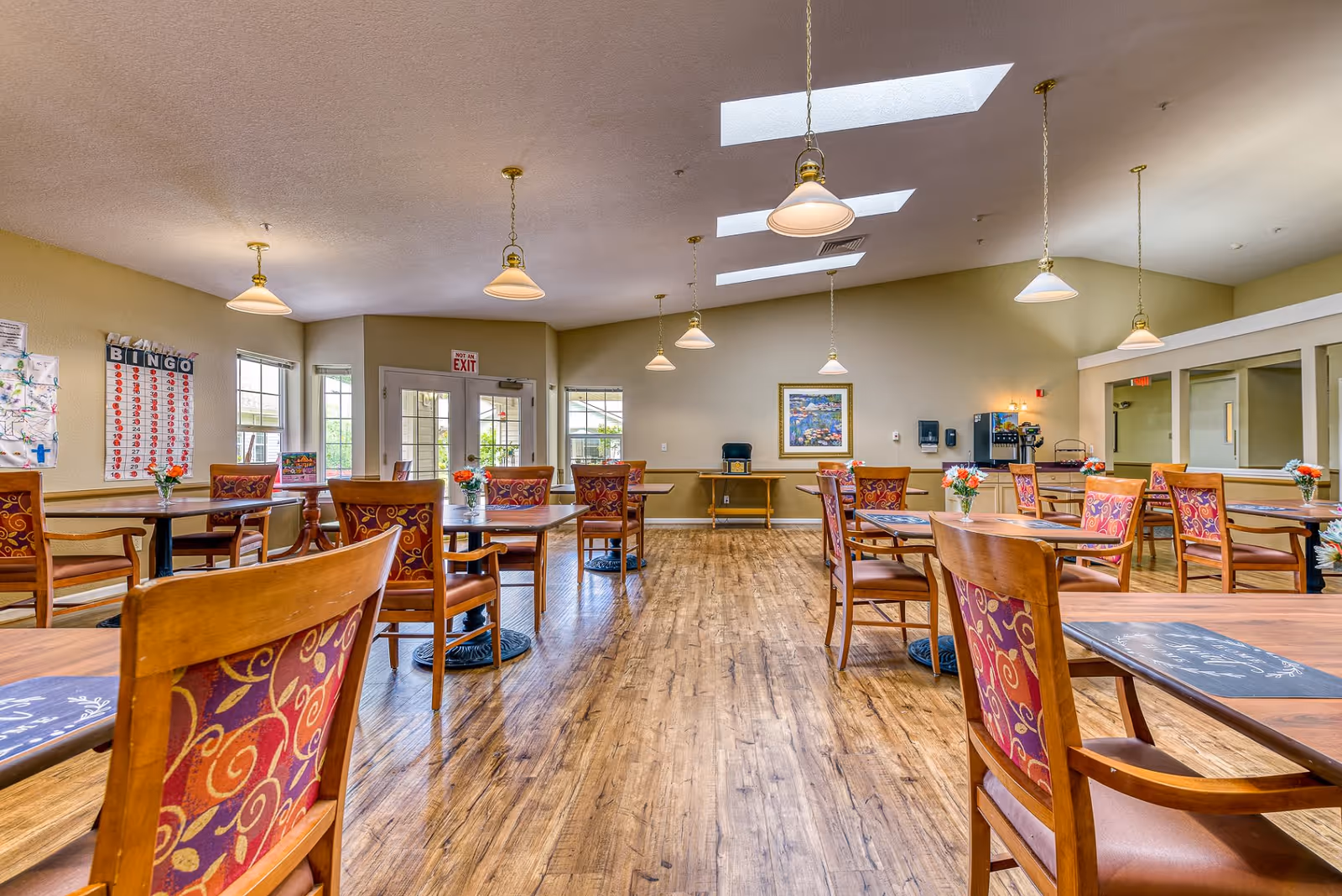 A bright and spacious dining room with wooden floors and multiple tables and chairs arranged neatly. Each table has a small vase with flowers. The room features hanging pendant lights and skylights in the ceiling, allowing natural light to fill the space. There is a bingo board on one wall and a coffee station in the back corner. Large windows and glass doors provide views of the outside.