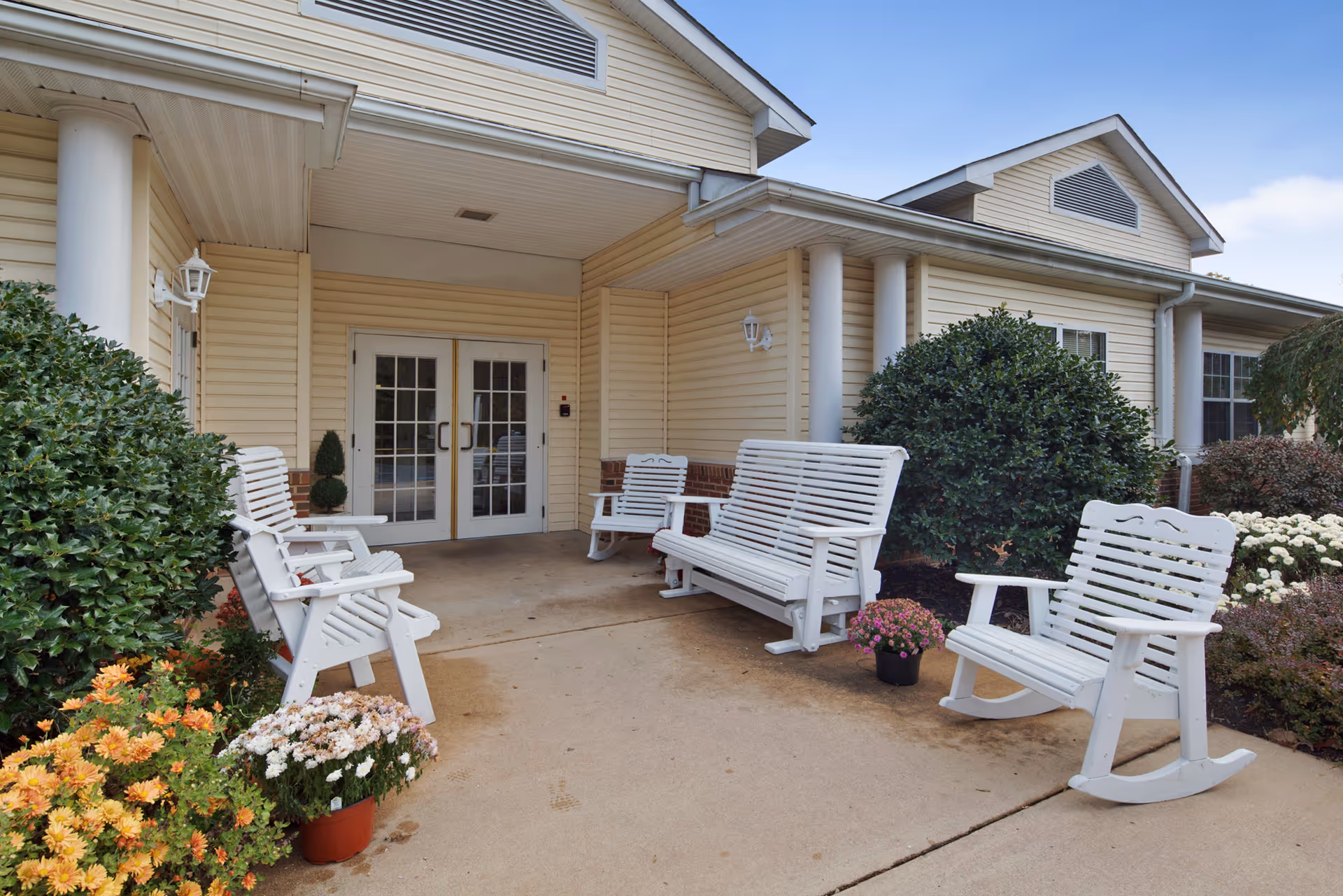 Covered front entrance of a senior living facility with white rocking chairs and benches, potted flowers, and shrubs.