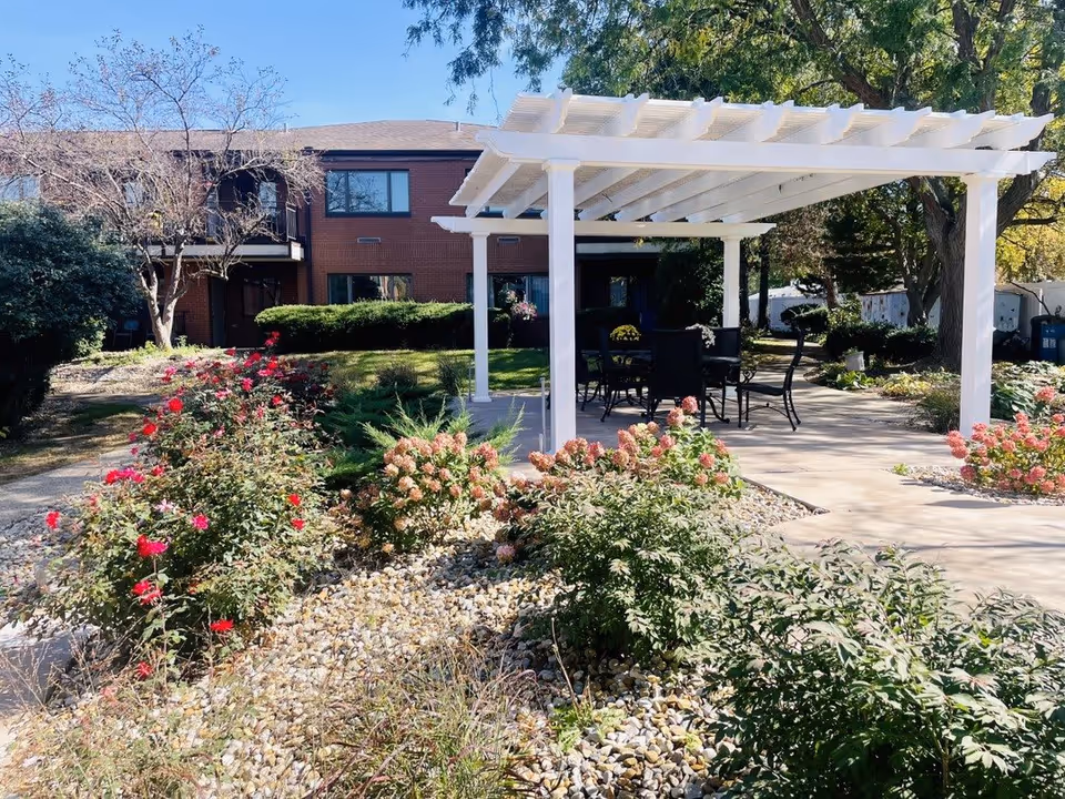 Outdoor garden area at Proctor Place Senior Living featuring a white pergola with black patio chairs and tables underneath. Surrounding the pergola are various flowering plants and bushes, with a two-story brick building in the background under a clear blue sky.
