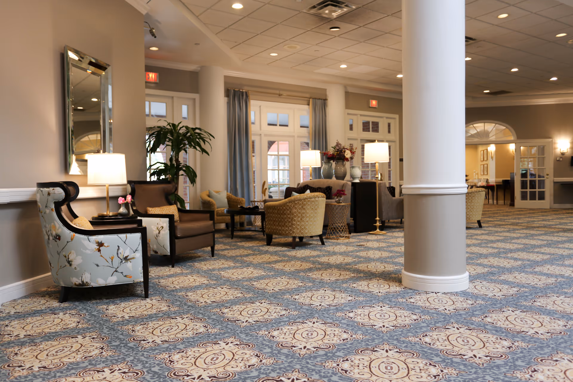 A spacious and well-lit common area with patterned carpet flooring, multiple seating arrangements including armchairs and sofas, side tables with lamps, decorative plants, and large windows with curtains allowing natural light to enter.