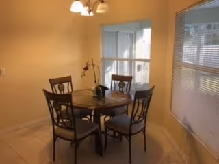 Small dining area with a round wooden table and four chairs under a chandelier next to windows with blinds.