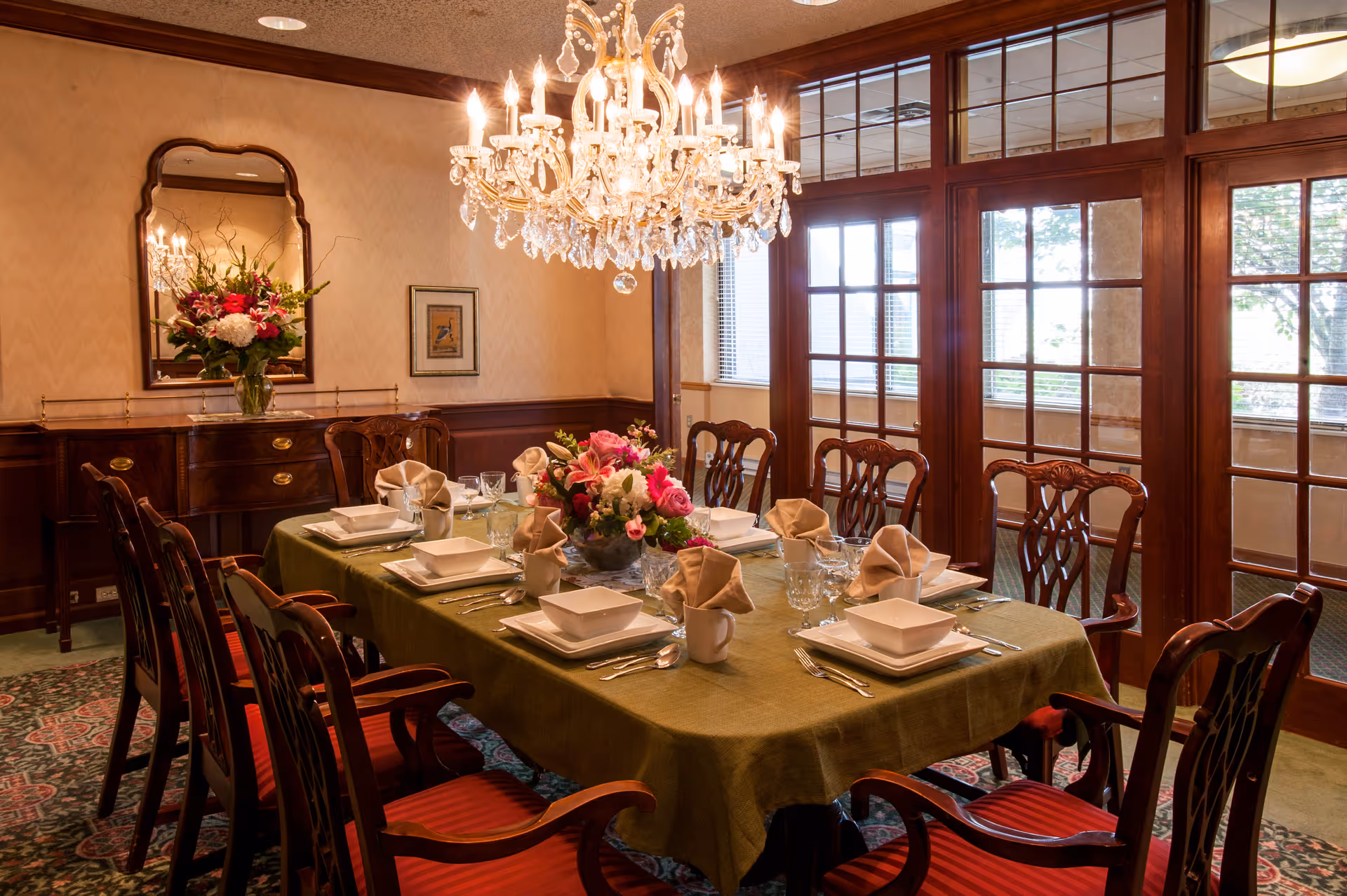Formal dining room with a chandelier, a table set for eight with floral centerpieces and wooden chairs in front of French doors.