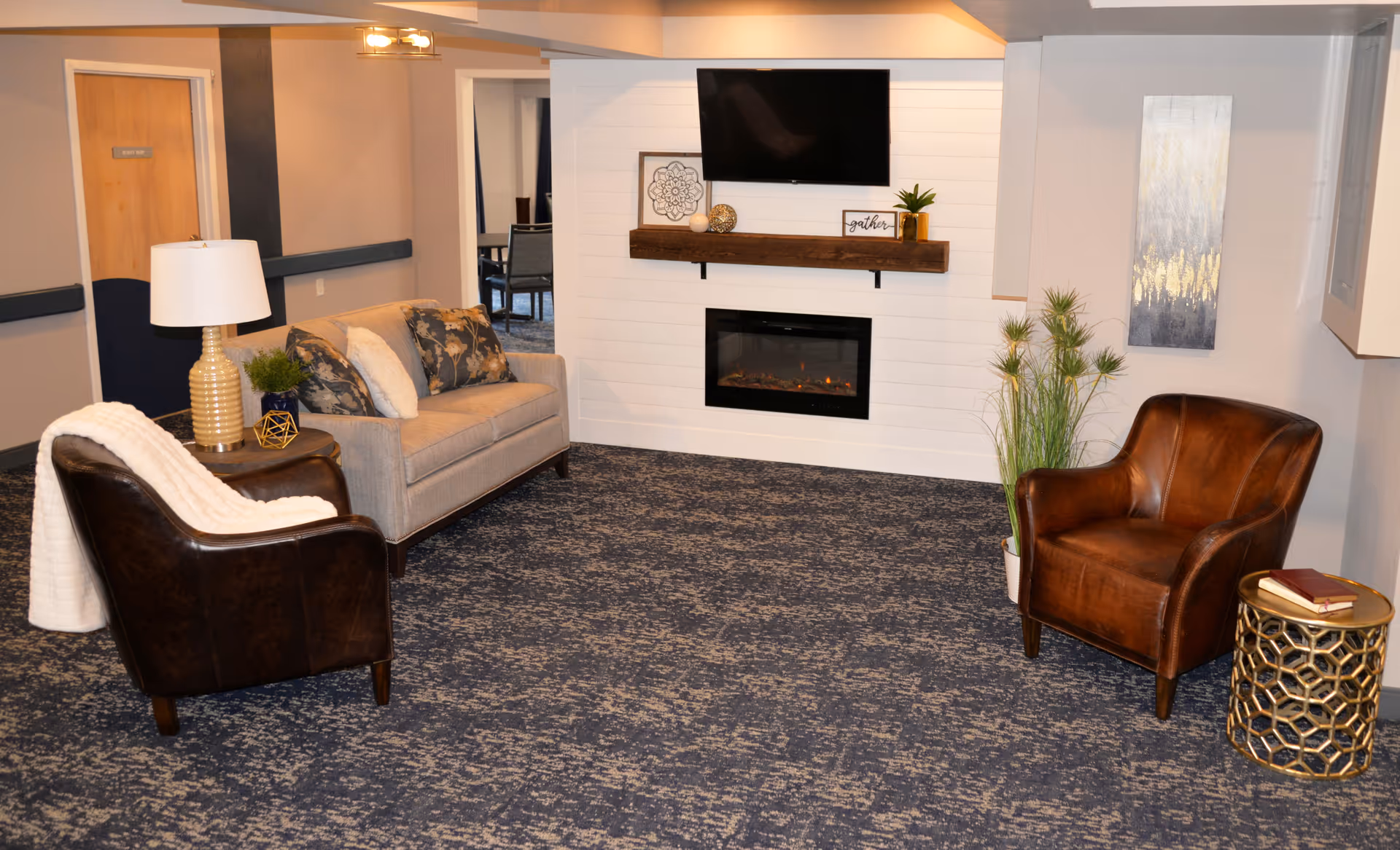 Seating area with a sofa and two leather armchairs facing a wall-mounted TV and electric fireplace in a decorated common room.