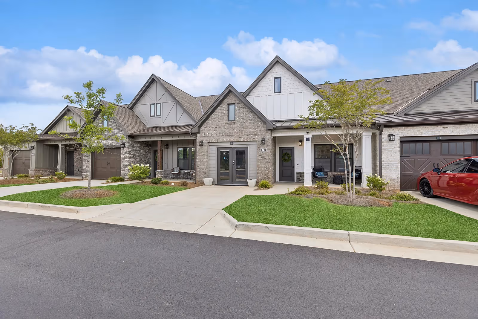 Exterior view of a modern senior living facility with multiple connected units featuring gabled roofs, brick and siding facades, small front porches with seating, manicured lawns, young trees, and a red car parked in front of one garage.