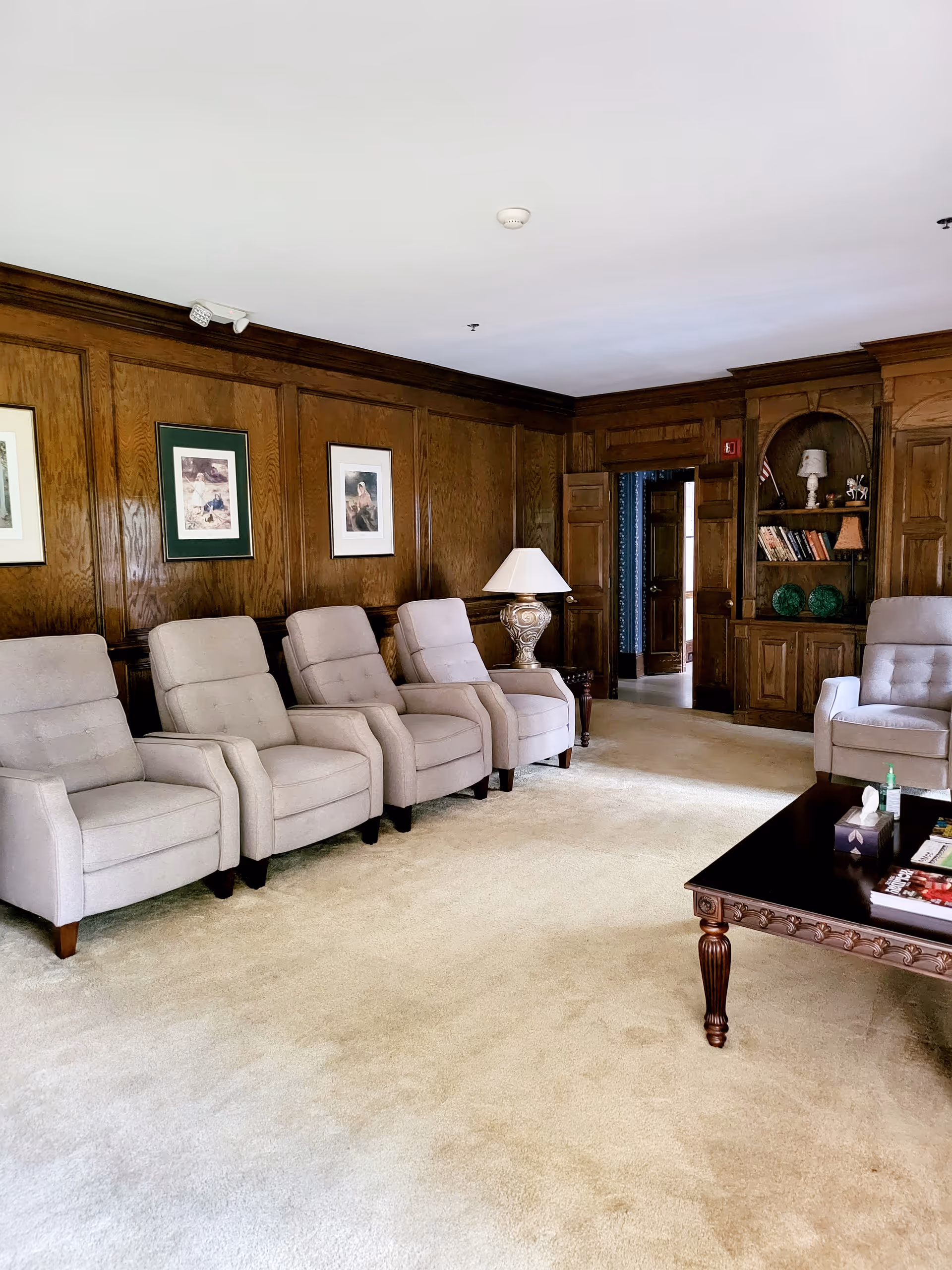 A cozy living room with five beige armchairs lined up against a wood-paneled wall adorned with framed artwork. A decorative table lamp sits on a small side table near an open doorway leading to another room. A wooden coffee table with magazines and a tissue box is positioned on a light carpeted floor. Built-in wooden shelves with books and decorative items are visible in the background.