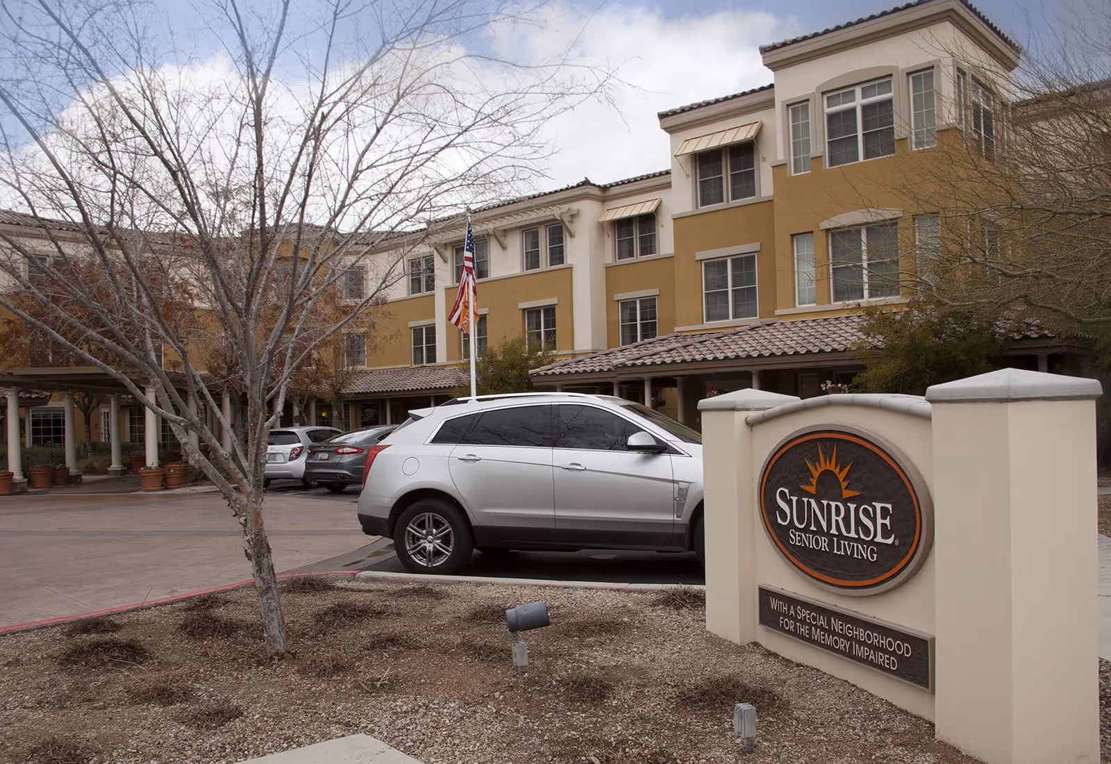 Exterior view of Sunrise Senior Living facility in Scottsdale, showing a multi-story building with beige and tan walls, several windows, a parked silver SUV, an American flag, and a sign in the foreground that reads 'Sunrise Senior Living with a special neighborhood for the memory impaired.'