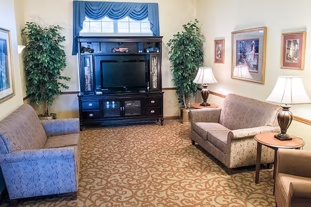 A cozy living room with two patterned sofas facing each other, a wooden entertainment center with a flat-screen TV, two potted plants on either side of the TV, framed artwork on the walls, and two table lamps on side tables providing warm lighting.