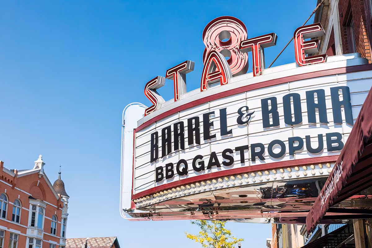 Retro theater marquee reading 'STATE' and advertising 'BARREL & BOARD BBQ GASTROPUB' over a downtown street against a clear blue sky.
