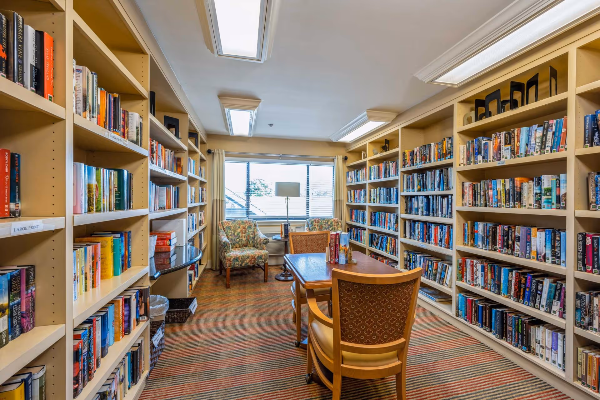 A cozy library room with bookshelves filled with books lining both sides of the room. There is a wooden table with two chairs in the center and two floral armchairs near a large window at the far end. The room is well-lit with ceiling lights and natural light from the window.