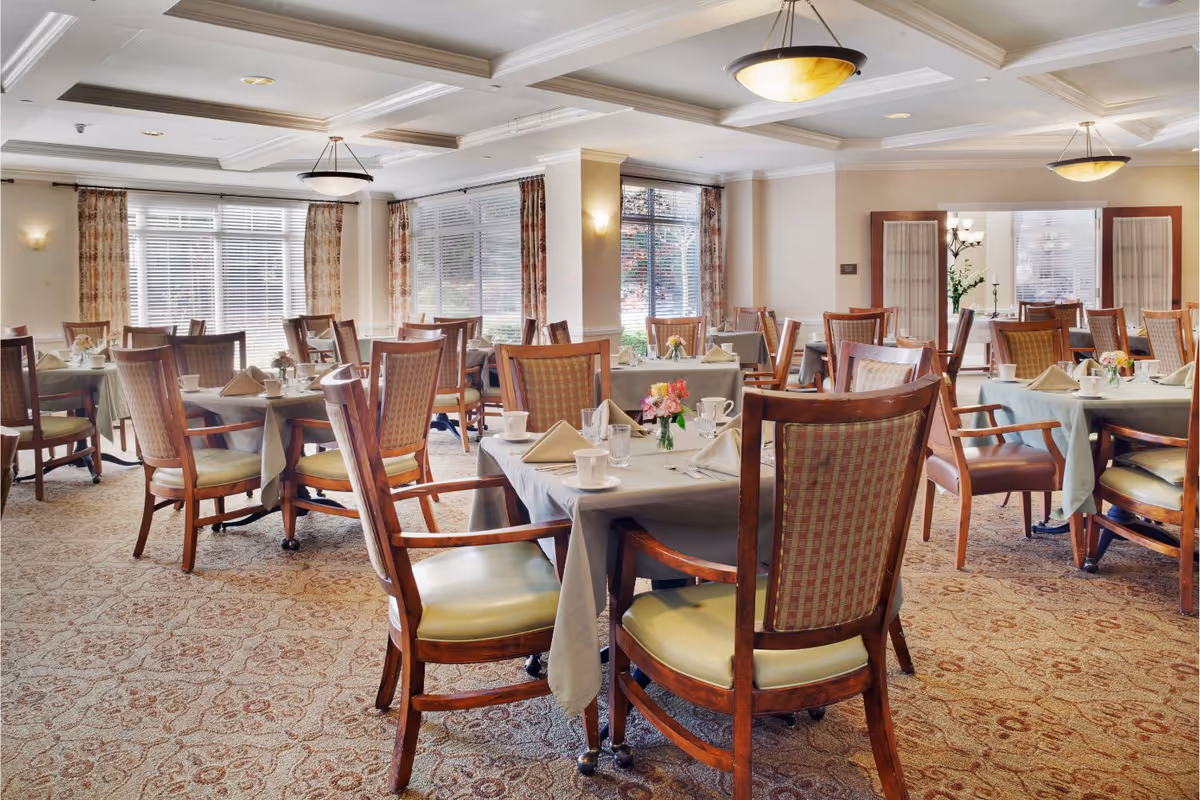 A bright and spacious dining room with multiple tables covered in gray tablecloths, each set with cups, glasses, napkins, and small floral centerpieces. The room features large windows with blinds and patterned curtains, wooden chairs with cushioned seats, and elegant ceiling light fixtures.