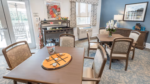 A bright and cozy common area in a senior living facility with two wooden tables surrounded by cushioned chairs. One table has a tic-tac-toe game board on it, and the other has a vase with flowers. The room features a window with patterned curtains, a sideboard with decorative items, a lamp, and a framed picture on the wall. A door leads to an outdoor patio area.
