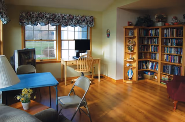 A cozy room with wooden flooring featuring a desk with a computer and wooden chair near a window with floral valance curtains. There is a blue table with a potted plant and two folding chairs, a bookshelf filled with books and decorative items, and a red armchair partially visible on the right.
