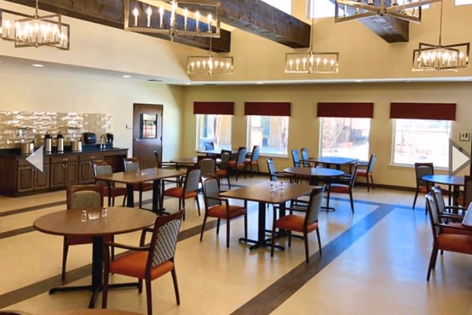 Spacious dining room with round and rectangular tables and chairs, chandeliers, a buffet counter, and large windows.