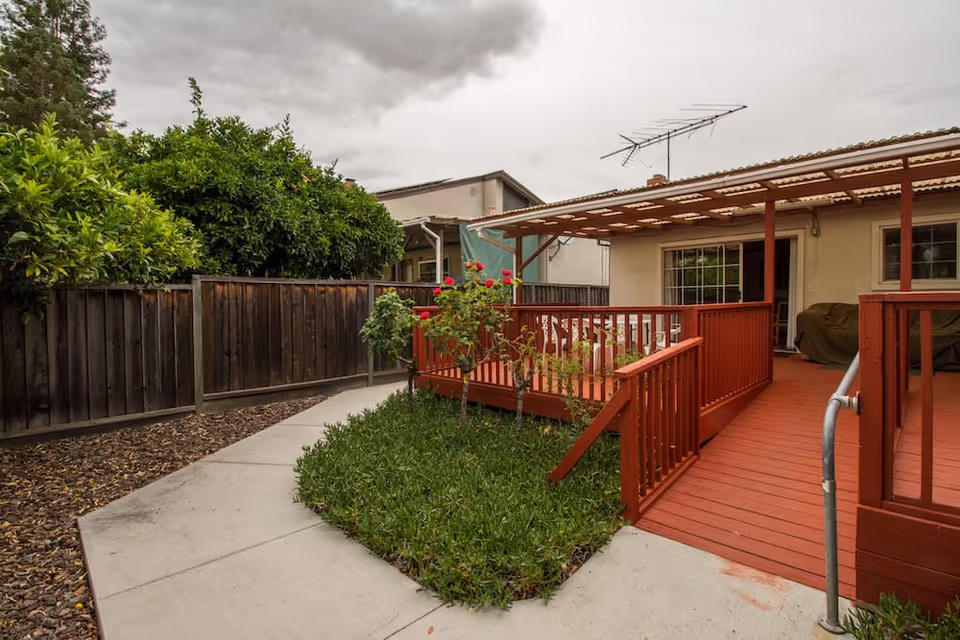 Backyard showing a red wooden accessible ramp and deck leading to a covered patio beside a lawn, concrete path, and wooden fence.