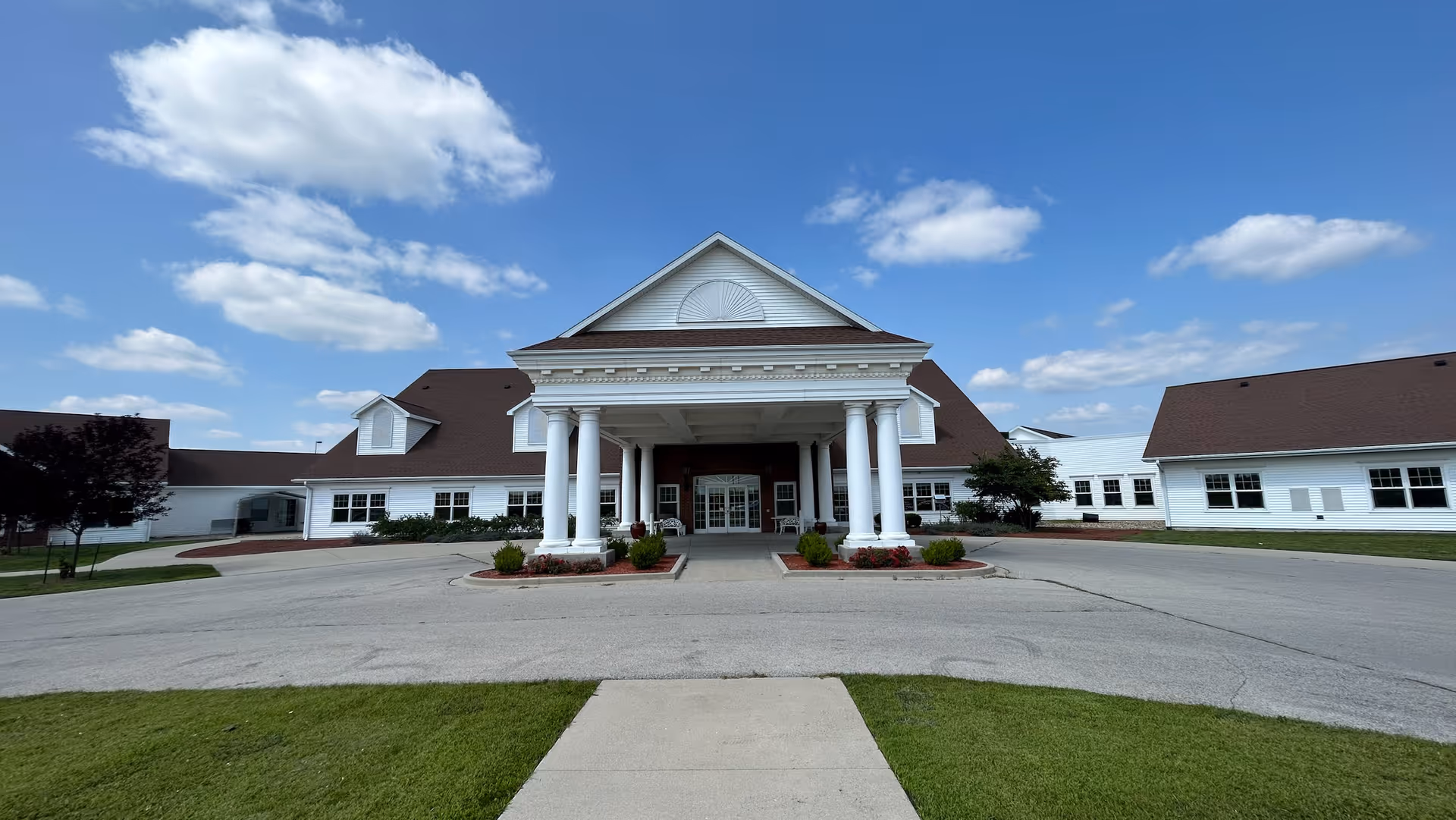 Front exterior view of a large white building with a covered entrance supported by six white columns, surrounded by a driveway and green lawn under a partly cloudy blue sky.