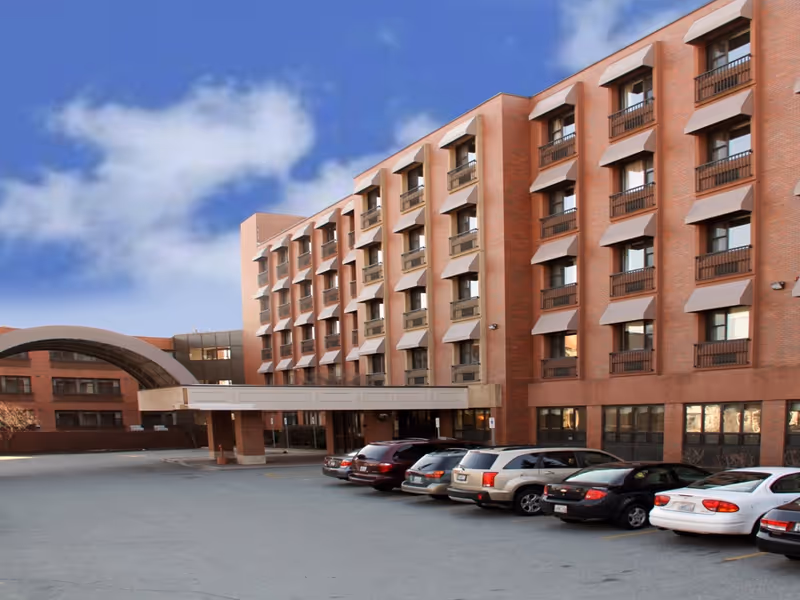 Exterior view of a multi-story brick building with several windows and a covered entrance. There are multiple cars parked in front of the building under a clear blue sky with some clouds.
