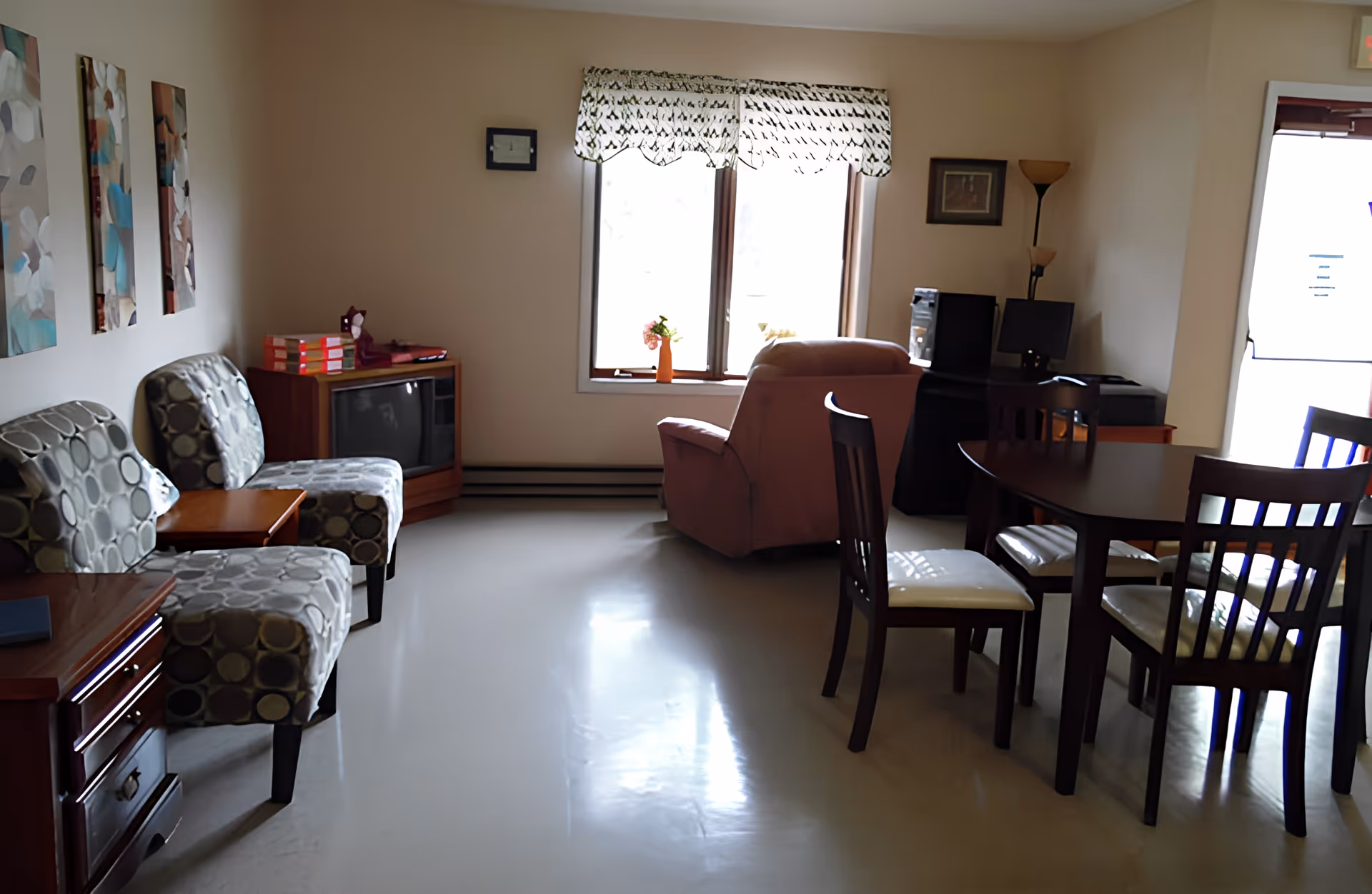 A cozy living room area with two patterned chairs and a small wooden table on the left, a vintage television on a wooden stand in the corner, a large window with a patterned valance letting in natural light, a comfortable armchair facing the window, a computer desk with a monitor and tower, and a dark wooden dining table with four chairs on the right side near a glass door.