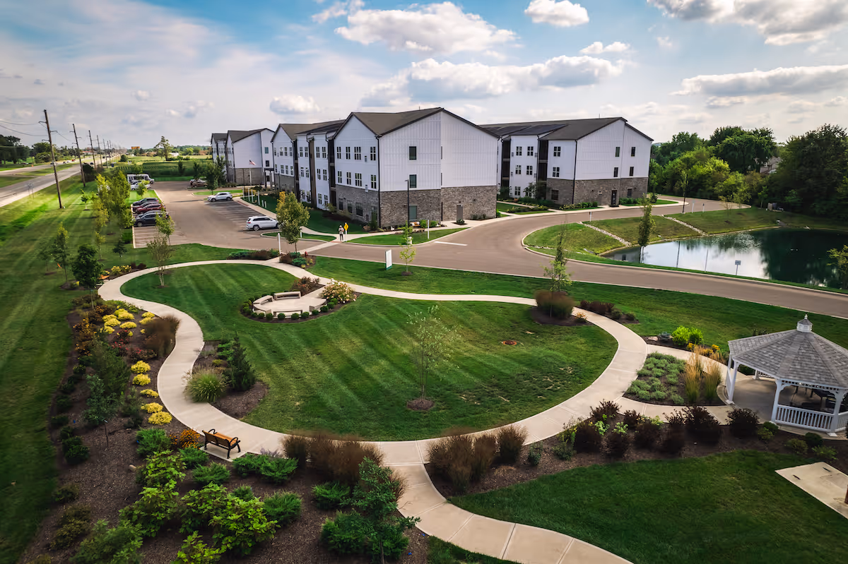 Aerial view of a senior living facility named Astral at Franklin showing a large, well-maintained green lawn with curved walking paths, landscaped gardens, a gazebo, a pond, and a three-story residential building in the background under a partly cloudy sky.