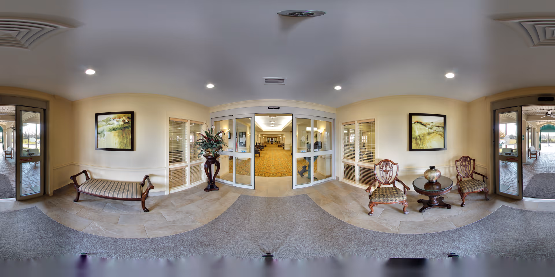 Entrance area of Oakmont Sterling Senior Community featuring double glass doors leading into a hallway with carpeted flooring. On either side of the doors are windows with blinds. To the left, there is a wooden bench with a cushioned seat beneath a framed landscape painting. To the right, there are two wooden armchairs with patterned cushions and a round wooden table with a decorative vase on top, also beneath a framed landscape painting. The floor near the entrance is tiled, transitioning to carpet inside. The ceiling has recessed lighting and air vents.