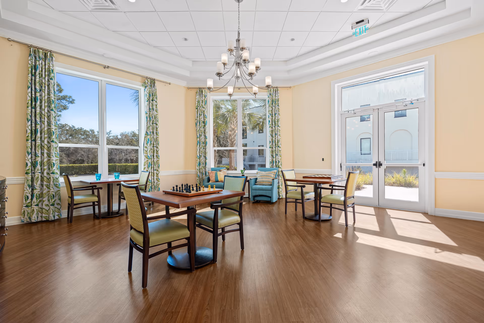 A bright and spacious common room with large windows and glass doors letting in natural light. The room features several small tables with chairs, one table has a chessboard set up. There are two blue armchairs with patterned cushions near the windows, and green leafy patterned curtains frame the windows. The floor is wooden, and a modern chandelier hangs from the ceiling.