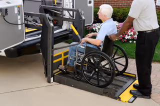 An elderly person in a wheelchair is being assisted by a caregiver while using a wheelchair lift attached to a vehicle outside a building with flowers and greenery in the background.