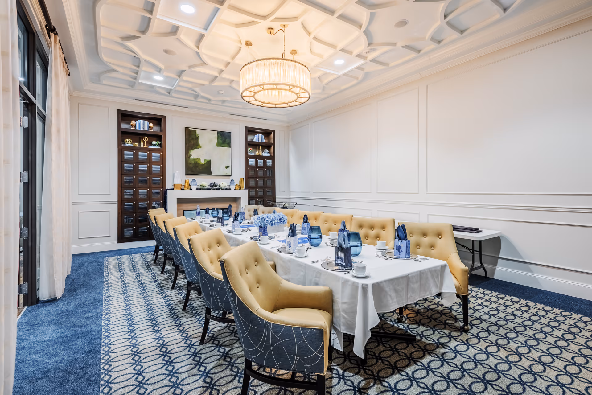 A formal dining room with a long rectangular table set for a meal. The table is covered with a white tablecloth and has blue glassware, white cups, and napkins arranged neatly. The chairs around the table are upholstered in a combination of yellow and blue fabric. The room features a patterned carpet, white paneled walls, a decorative ceiling with recessed lighting, and a large circular chandelier. There are built-in shelves and a fireplace at the far end of the room.