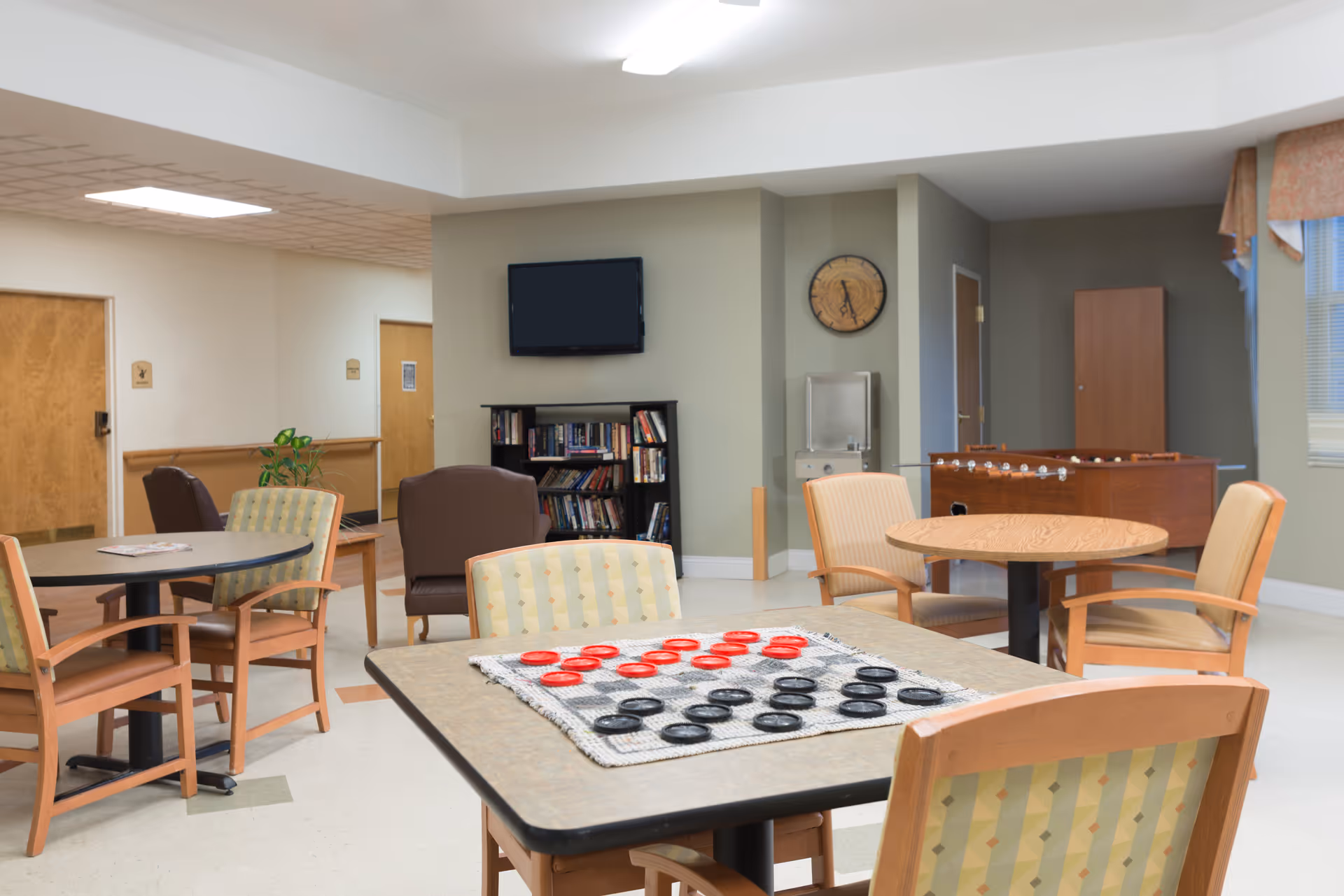 A common area in a senior living facility with several tables and chairs arranged for social activities. One table has a checkers game set up with red and black pieces. In the background, there is a bookshelf filled with books, a wall-mounted TV, a clock, a water fountain, and a foosball table near a window with curtains.
