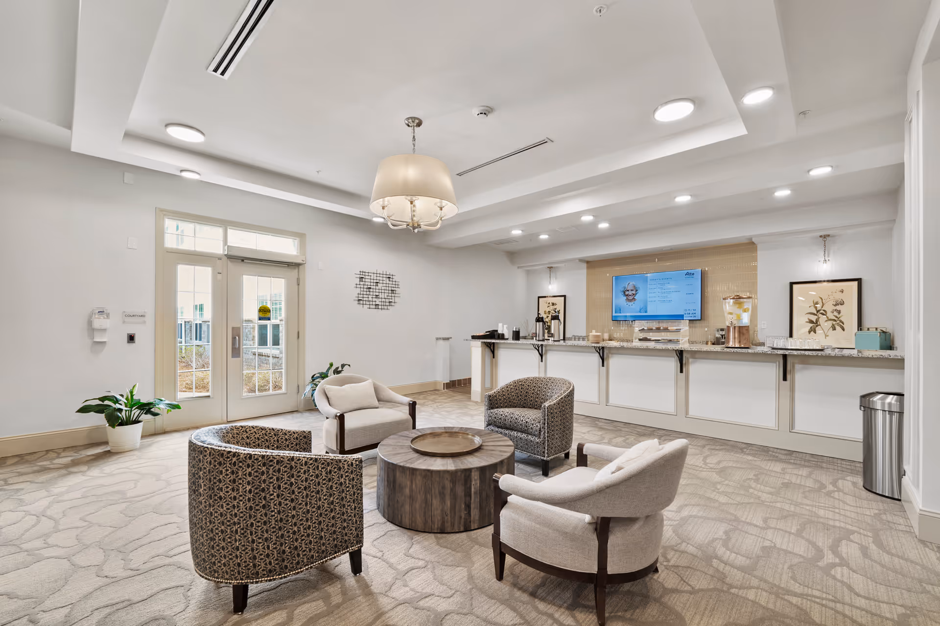A bright and spacious common area with four upholstered chairs arranged around a round wooden coffee table. The room features a large counter with a granite top, a mounted flat-screen TV displaying information, decorative framed artwork on the walls, and a modern chandelier hanging from the ceiling. There are double glass doors leading outside, potted plants, and soft carpeting with a subtle pattern.