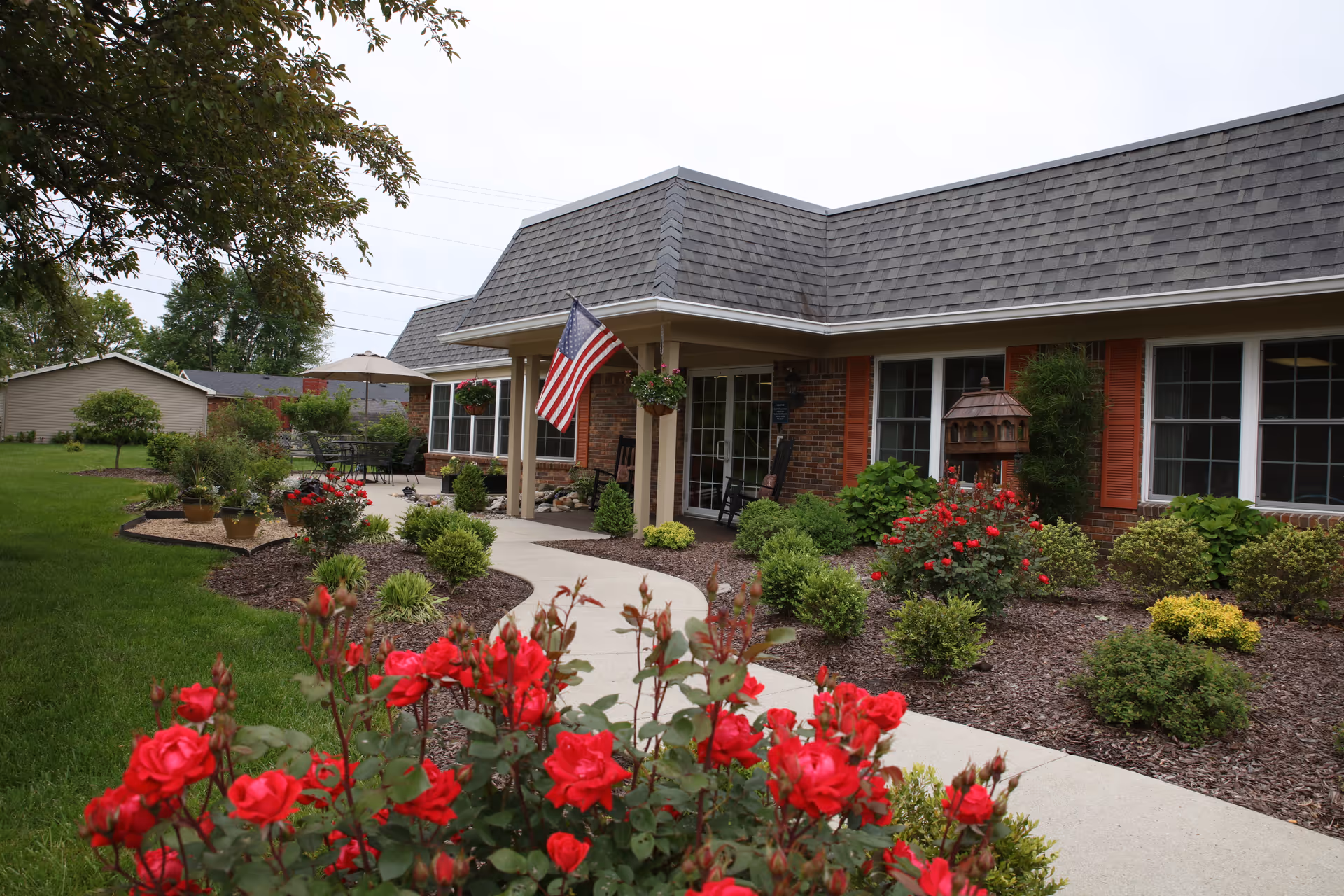Front entrance of a brick senior living building with an American flag, landscaped garden, and red roses along a curved walkway.