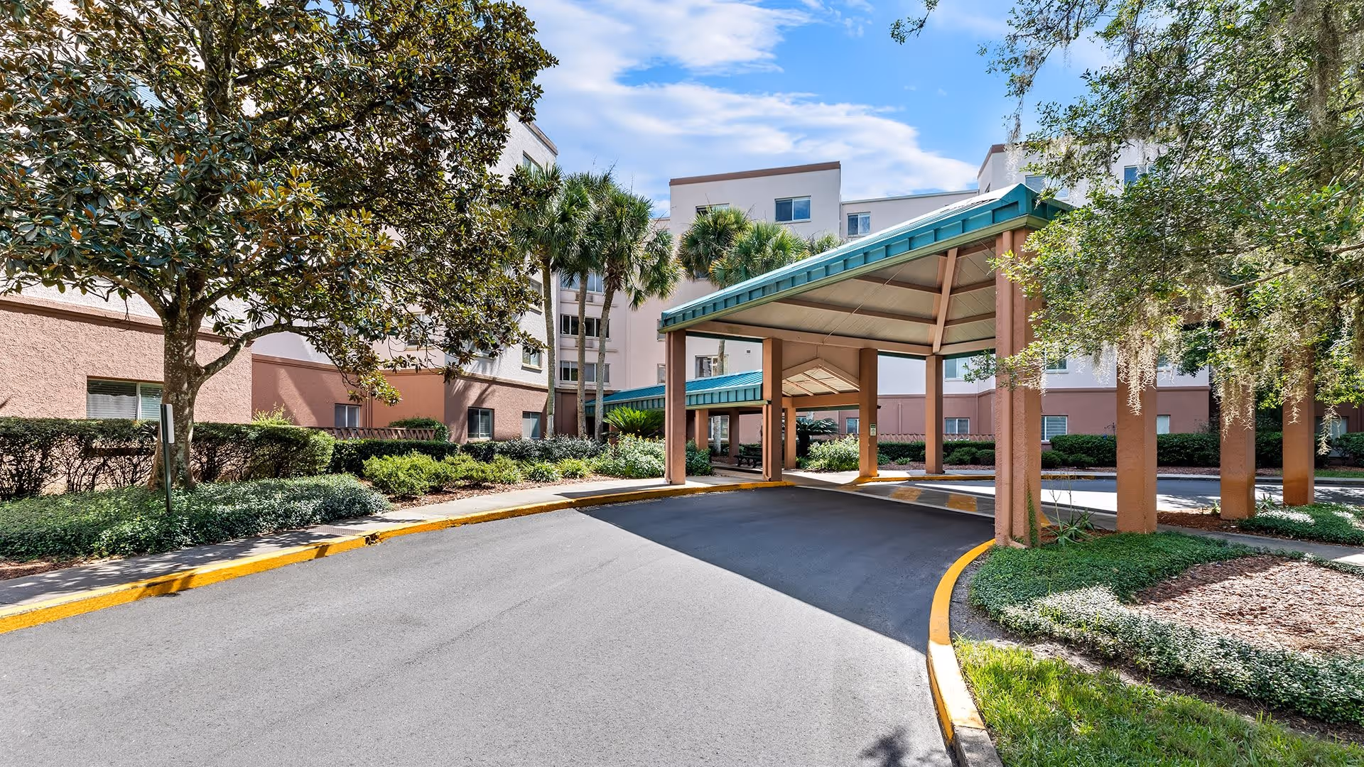 Entrance driveway of Atrium at Gainesville senior living facility with a covered drop-off area, surrounded by landscaped greenery including palm trees and bushes, under a partly cloudy blue sky.