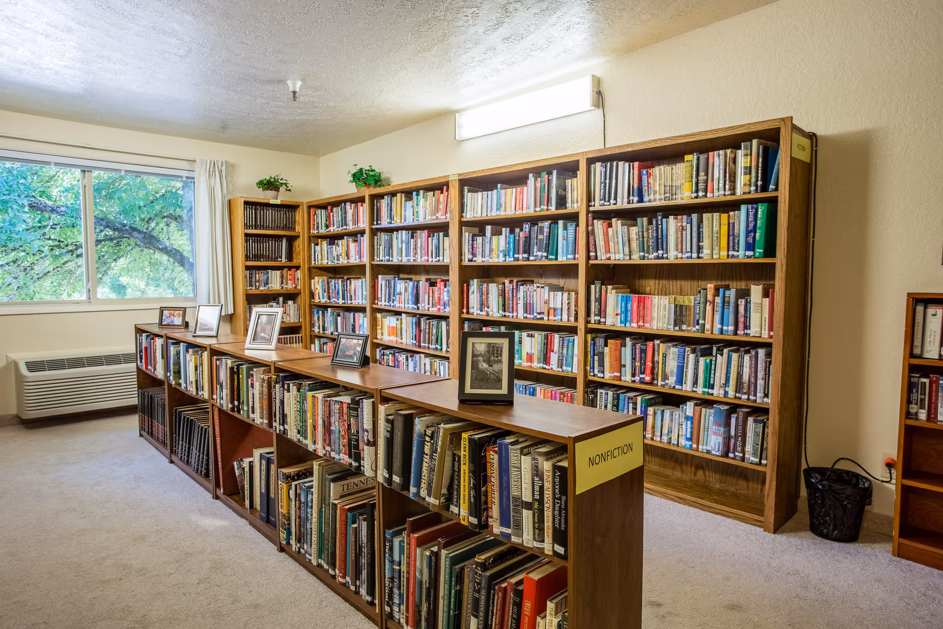 Interior view of a library room with wooden bookshelves filled with books. The shelves are labeled with categories such as 'NONFICTION' and 'FICTION'. There are framed photos displayed on top of the shelves. A window with a view of green trees is on the left side, and a wall-mounted light fixture illuminates the room.