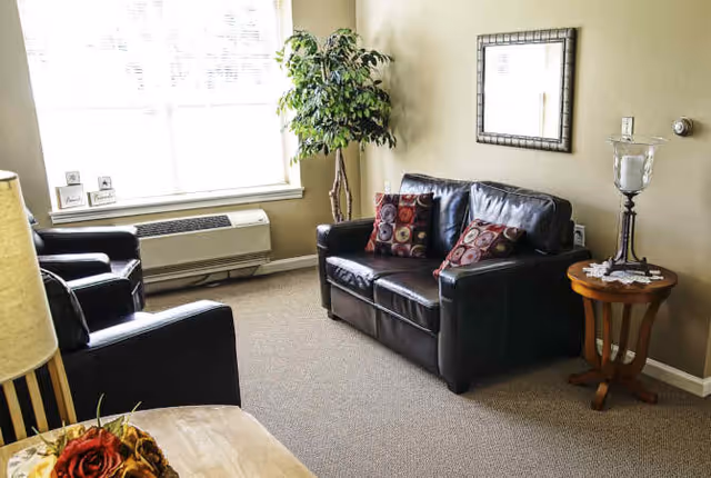 A cozy living room area with a black leather loveseat and two matching armchairs. The loveseat has two decorative pillows with circular patterns. There is a wooden side table with a large candle holder and a doily on it. A large window with blinds lets in natural light, and a tall green plant is placed near the window. A framed mirror hangs on the beige wall above the loveseat.